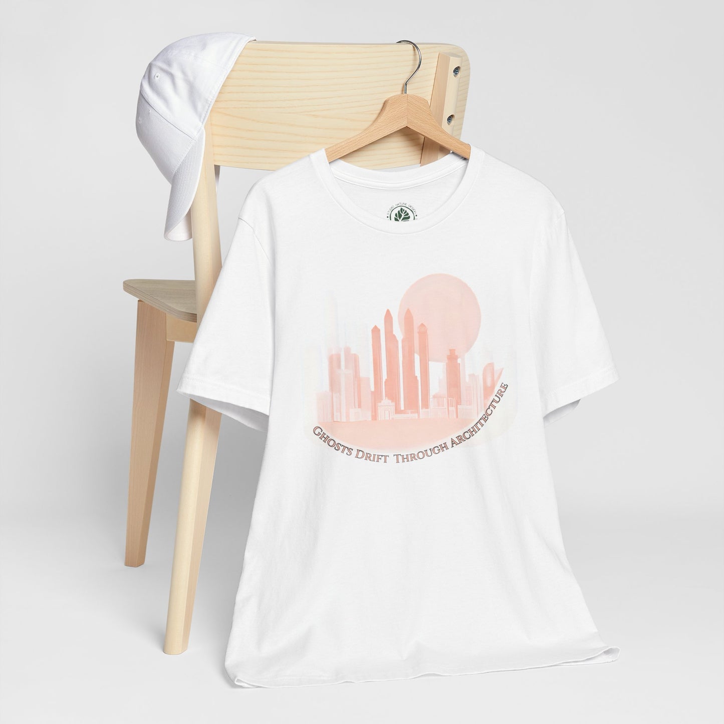 White unisex t-shirt featuring a coral city skyline with the text “Ghosts Drift Through Architecture” beneath, symbolizing poetic urban design on a wooden chair against a white background.