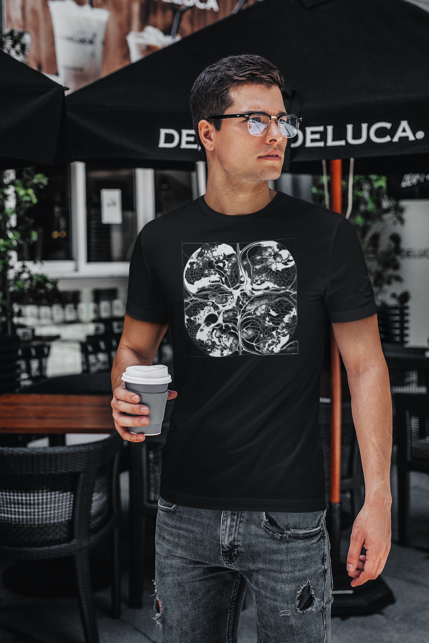 A person standing near outdoor café tables, wearing a black T-shirt with a white city-meets-neuron line art design and holding a coffee cup.