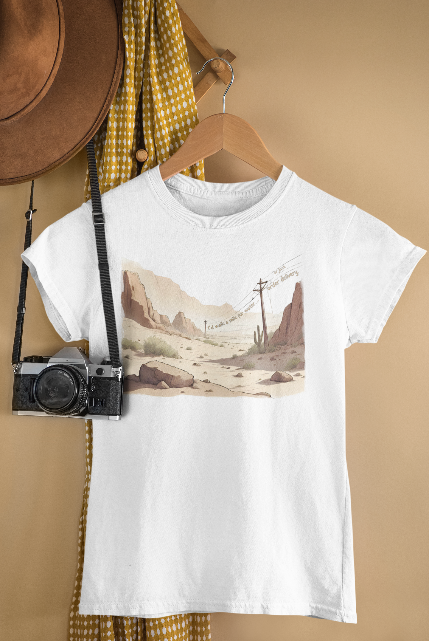 White t-shirt with a desert landscape, telegraph poles and witty caption “I’d walk a mile for water… or just order delivery.” design hanging on a wooden hanger with a camera and hat in the background.