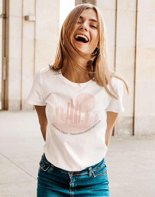 Woman wearing a white t-shirt with text, standing indoors. Unisex white t-shirt featuring a coral city skyline with the text “Ghosts Drift Through Architecture” beneath, symbolizing poetic urban design.
