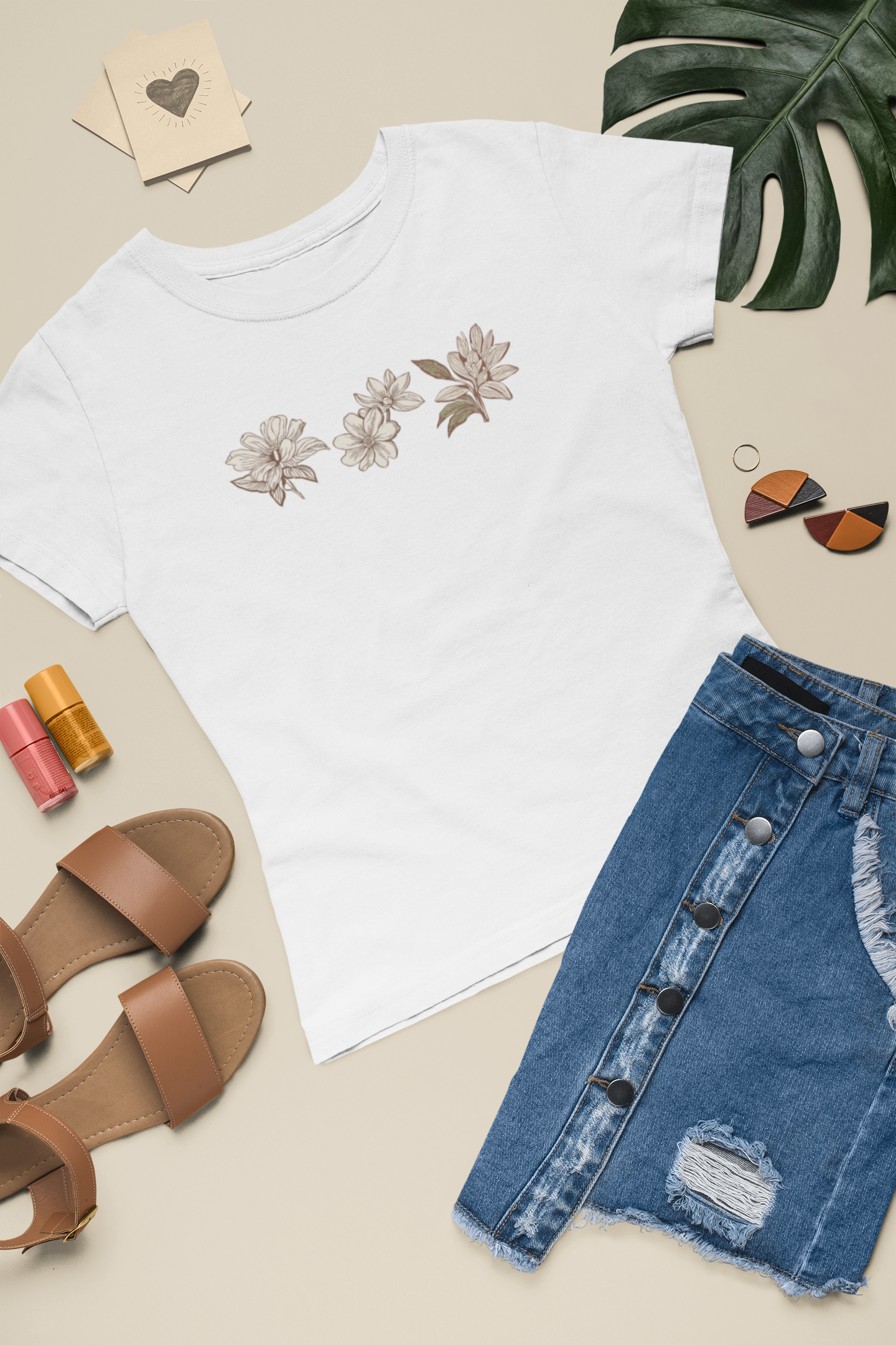 White t-shirt sustainable fashion floral tee with eco-friendly organic cotton, denim skirt, sandals, nail polish, and a plant on a beige surface.
