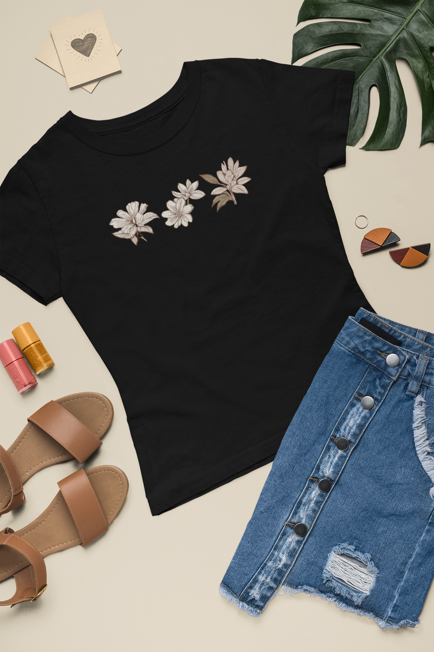 Black t-shirt sustainable fashion floral tee with eco-friendly organic cotton, denim skirt, sandals, nail polish, and a plant on a beige surface.