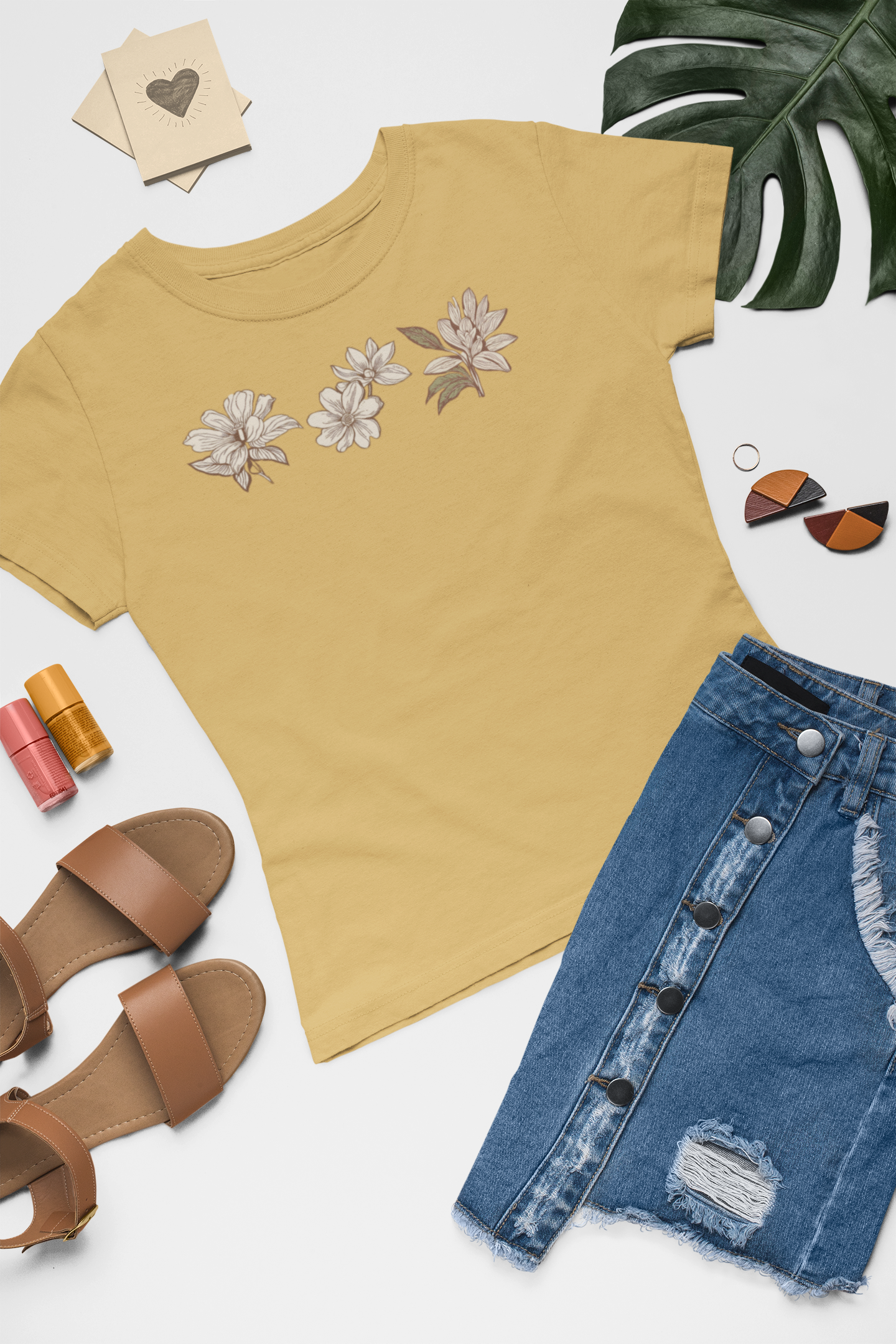 Mustard t-shirt sustainable fashion floral tee with eco-friendly organic cotton, denim skirt, sandals, nail polish, and a plant on a beige surface.